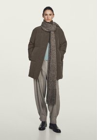 Kaki pufferjakke med høj krave, en blå sweater underneath, grå brede bukser og et tekstureret, frynset tørklæde i neutrale nuancer.