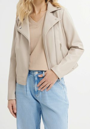 Woman wearing beige leather biker jacket over tan v-neck shirt and light blue high-waisted jeans, hand resting on waistband.