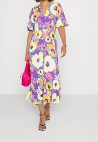 Abito maxi floreale in viola con accenti gialli, rosa e verdi. Presenta un scollo a V, maniche corte a volant e uno spacco alta fino al ginocchio.