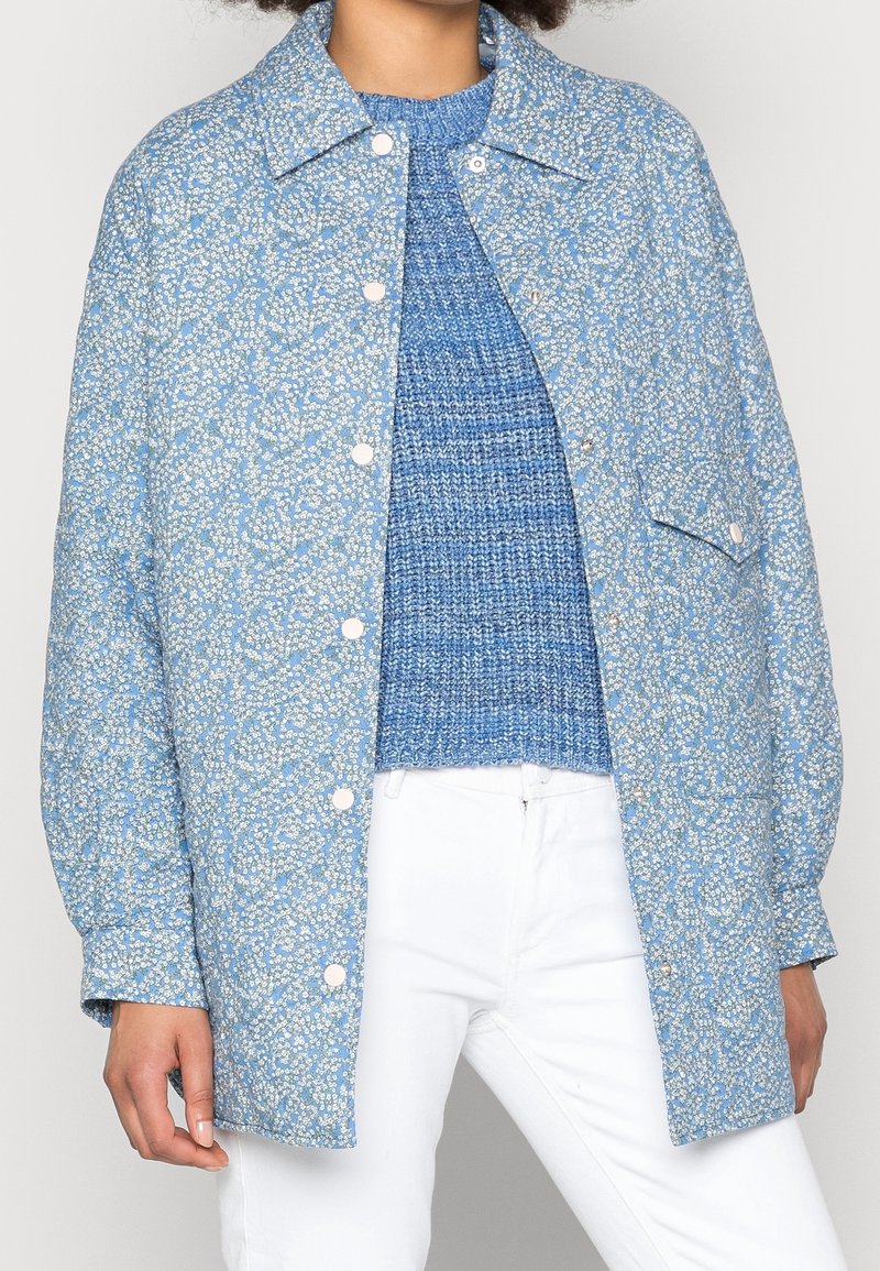 Gequiltte blauwe jas met een klein bloemenpatroon, enkele zak en drukknoopsluitingen. Draagt een gestructureerde blauwe trui en witte broek.
