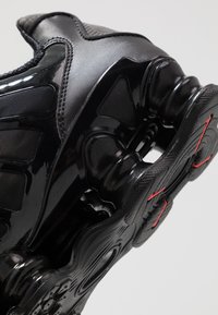 Μαύρο αθλητικό παπούτσι με γυαλιστερό συνθετικό υλικό, υφασμάτινες λεπτομέρειες και ορατή καμπυλωτή μαύρη σόλα με κόκκινα σημεία.