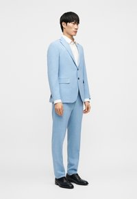 Ανοιχτό μπλε κοστούμι από υφασμάτινη ύφανση με υφή· μονόπετο σακάκι, γιακάδες με εγκοπή και παντελόνια που ταιριάζουν. Μαύρα παπούτσια ολοκληρώνουν το σύνολο.