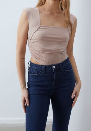 Beige, tætsiddende top med firkantet halsudskæring og rynket sideffekt, kombineret med mørkeblå skinny jeans. Glat stof, minimalistisk design.