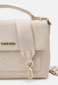 Beige crossbody taška s texturovanou koženkou, s embosovaným logem, nastavitelným popruhem a zlatými akcenty.