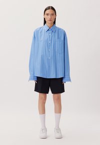 Camicia oversize a righe blu con chiusura a bottoni e tasca, abbinata a pantaloni corti neri e calze bianche, su uno sfondo neutro.