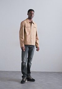 Tan lightweight jacket with large front pockets and button closure, paired with dark jeans and black sneakers. Smooth texture, relaxed fit.