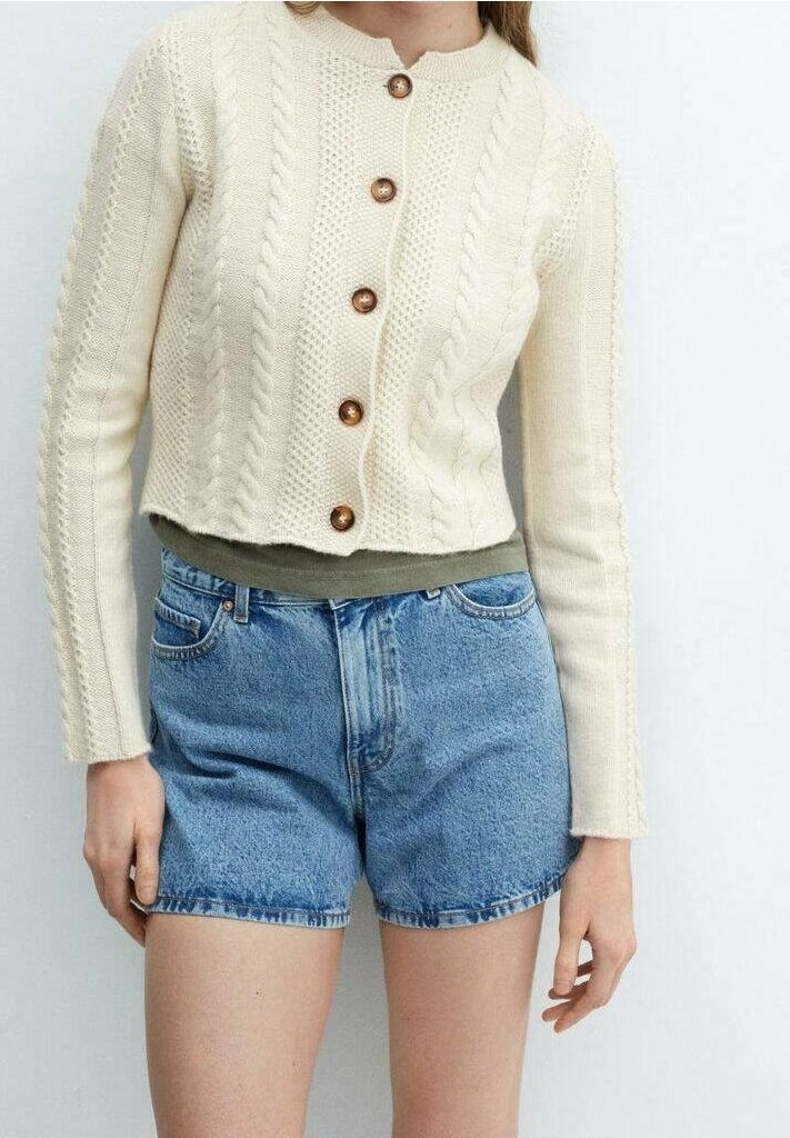 Person in einer cremefarbenen, gestrickten, kurzen Strickjacke mit Holzknöpfen, grünem Oberteil und hochgeschnittenen blauen Denim-Shorts.