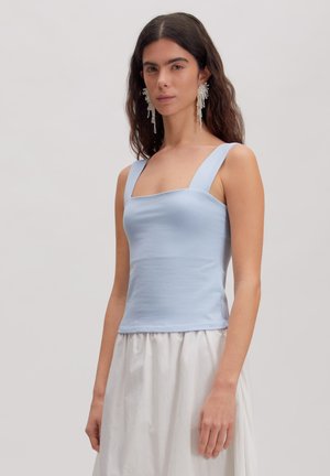 Woman with long dark hair wears sleeveless light blue top and white skirt, paired with large dangling pearl earrings.