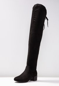 Bota negra por encima de la rodilla hecha de ante, con punta redondeada y un tacón de bloque texturizado, que presenta un detalle de cordones en la parte posterior.