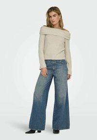 Suéter de punto beige sin hombros con textura acanalada, combinado con jeans de mezclilla azul de pierna ancha y botas negras hasta el tobillo.
