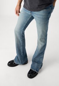 Jeans acampanados de color azul claro en tejido de mezclilla con un ligero desgaste, con un diseño clásico de cinco bolsillos y combinados con zapatos negros sin cordones.