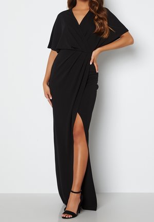 Femme portant une robe de soirée noire style portefeuille avec manches courtes et une fente haute à la cuisse, assortie à des talons hauts noirs à bride de cheville.