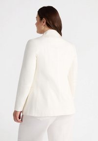 Biała marynarka o dopasowanym kroju, gładkiej fakturze materiału, z klapami w kształcie litery V i dopasowanym tyłem. Posiada boczne kieszenie i minimalne detale szycia.