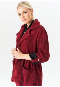 Red knit jacket with a shawl collar, black trim, two front pockets, adjustable drawstring waist, and textured pattern.