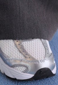 Baskets de course en maille blanche avec accents argentés, surface texturée et semelle en caoutchouc noir. Partiellement couvertes par le pantalon en tissu gris.