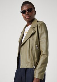 Jachetă din piele de culoare verde măslinie, de tip biker, cu fermoare și accesorii din argint. Are un design ușor ajustat, cu mâneci lungi și guler cu rever.