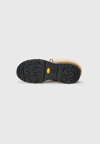 Visning av en brun boots svart gummisåle, med et teksturert mønster og en gul logo på hælen, satt mot en hvit bakgrunn.