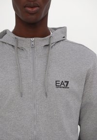 Siva zip-up majica napravljena od mekog materijala, sa kapuljačom koja se može podešavati i crnim izvezenim logom na lijevoj strani grudnog koša.