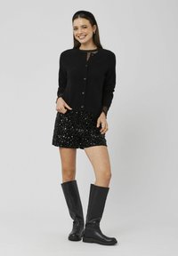 Cardigan noir en maille avec boutons, associé à une jupe noire à sequins et des bottes noires jusqu'aux genoux. Texture lisse et accents brillants.