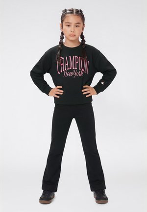 Ragazza con capelli intrecciati che indossa una felpa nera Champion e pantaloni, in piedi con sicurezza con le mani sui fianchi su uno sfondo bianco.