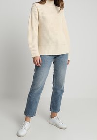 Frau trägt einen cremefarbenen Rollkragenpullover, hellblaue gerade Jeans und weiße Sneaker vor einem schlichten hellen Hintergrund.