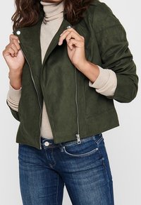 Veste courte en daim vert foncé avec fermeture éclair, boutons-pression et poches latérales, portée sur un col roulé beige et un jean bleu.