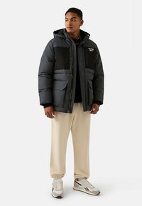 Schwarze und graue Steppjacke mit Kapuze, verfügt über Fronttaschen und Reißverschlusstaschen auf der Brust, getragen über einem schwarzen Oberteil und hellbeigen Hosen.