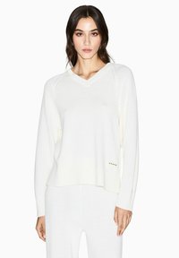 Cremefarbener V-Ausschnitt Pullover mit langen Ärmeln. Mit gerippten Bündchen und Saum sowie einem dezenten Logo an der Seite. Strickstruktur.