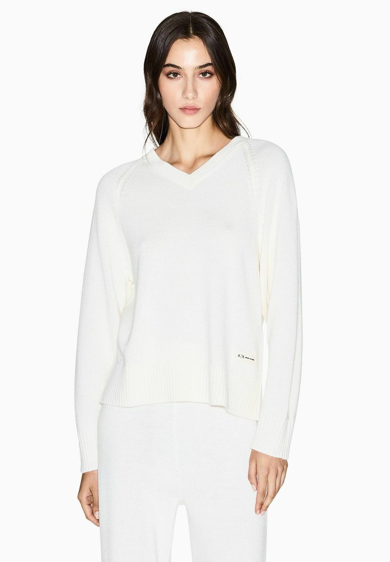 Cremefarbener V-Ausschnitt Pullover mit langen Ärmeln. Mit gerippten Bündchen und Saum sowie einem dezenten Logo an der Seite. Strickstruktur.