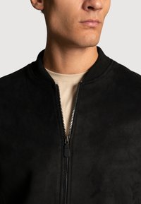 Veste bomber en daim noir avec un col côtelé et une fermeture éclair à l'avant. Présente une texture lisse et une silhouette ajustée.
