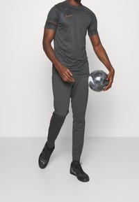 T-paita tummanharmaata urheiluvaatetta oransseilla yksityiskohdilla, yhdistettynä harmaisiin housuihin ja mustiin urheilukenkiin. Henkilö pitää jalkapalloa.