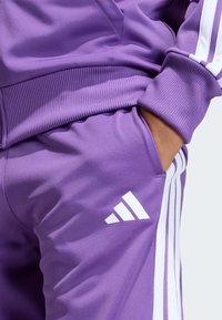 Violetti urheilupuku, jossa on resorihihansuut, valkoiset sivuraidat ja valkoinen logo lahkeessa. Sileä, joustava kangaspinta.