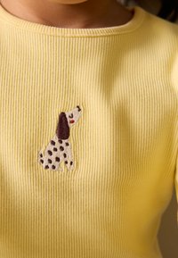 Žuta rebrasta majica sa smeđim i bijelim izvezenim psom s mrljama, smještenim na području prsa. Jednostavan okrugli izrez na vratu.