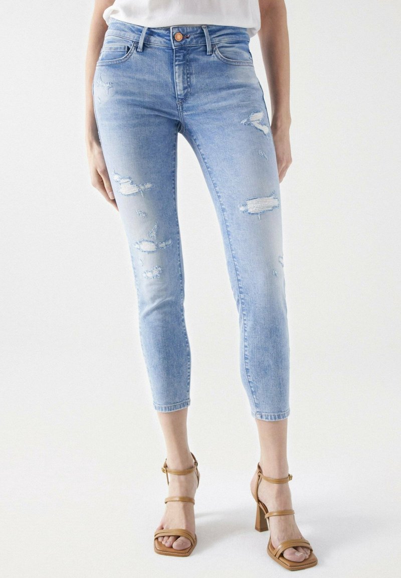 Femme portant un jean skinny bleu clair déchiré, raccourci au-dessus de la cheville, et des sandales à talons beiges avec des brides.