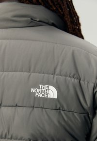 Grå pufferjakke med quiltet design, høy krage og en fremtredende hvit logo av "THE NORTH FACE" på baksiden.