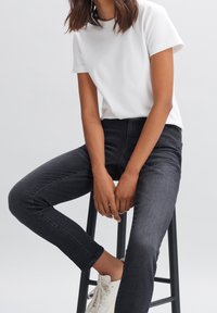 Personne portant un t-shirt blanc, un jean skinny noir et des baskets blanches, assise sur un tabouret noir devant un fond uni.