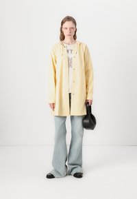 Chaqueta amarilla tipo botón con capucha, llevada sobre una camiseta gráfica, combinada con unos pantalones de mezclilla acampanados de color azul claro y zapatos planos negros. Sostiene un bolso negro.