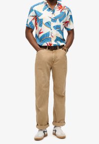 Camisa de manga corta y botones con un patrón de hojas colorido, combinada con pantalones de algodón beige y zapatillas blancas con detalles negros.