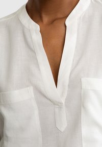 Blusa bianca in lino con colletto alla mandarino, caratterizzata da scollatura a V e due tasche frontali. Leggera e dal design minimalista.