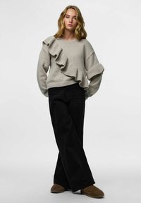 Svijetlosivi pleteni džemper s rebrastom teksturom i velikim volanom sa lijeve strane. Uparen s crnim trapericama širokih nogavica i smeđim cipelama.