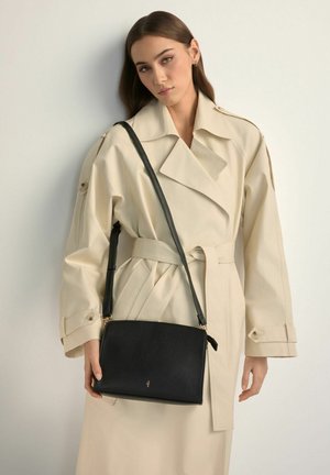 Kobieta w beżowym płaszczu trencz, niosąca czarną skórzaną torbę na ramię z długim paskiem na jednolitym jasnym tle.