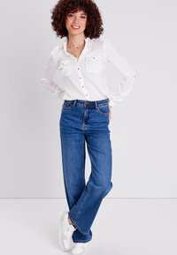 Camicia bianca a maniche lunghe con due tasche frontali, abbinata a jeans blu a vita alta e gamba dritta e scarpe da ginnastica bianche su uno sfondo neutro.