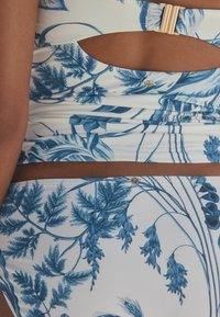 Blaue Bikini-Oberteil mit Blumenmuster und Ausschnitten, aus glattem Stoff gefertigt. Verfügt über einen goldenen Verschluss und dazu passende hochgeschnittene Hose.