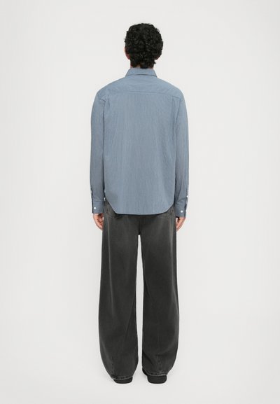 Homme aux cheveux bouclés de dos, portant une chemise à rayures bleues à manches longues et un pantalon noir à jambes larges sur fond blanc.
