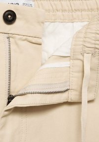 Pantalon beige avec fermeture éclair partiellement ouverte, bouton noir, et tissu de doublure intérieure blanche visible.