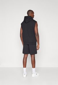 Haut à capuche sans manches noir, associé à un short noir. L'ensemble présente une coupe décontractée et est fabriqué dans un tissu lisse. Des baskets blanches complètent le look.
