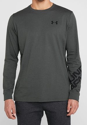 Mann trägt ein dunkelgraues langärmeliges Under Armour Shirt mit Logo auf der Brust und Branding-Text auf dem linken Ärmel, kombiniert mit dunkler Hose.