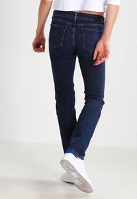 Mörkblå jeans med slim fit, med medelhög midja, två bakfickor och synlig sömnad. Bärs med vita sneakers.