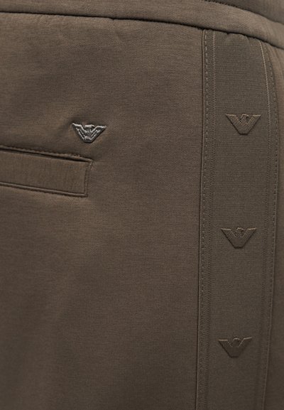 Κοντινή λήψη τσέπης καφέ παντελονιού με κάθετη λωρίδα που φέρει επαναλαμβανόμενα ανάγλυφα λογότυπα αετού κατά μήκος της πλευρικής ραφής.