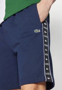 Man wearing navy blue Lacoste shorts with green crocodile logo and branded black and white side stripe, hand in pocket.